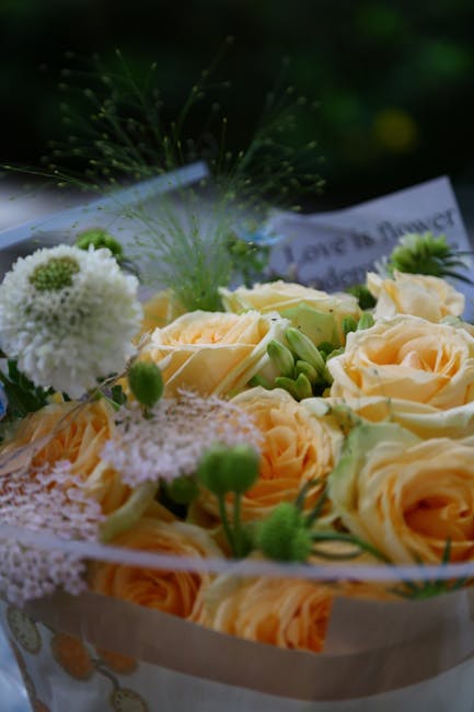 A close-up of a floral arrangement featuring soft peach-colored roses with full, slightly ruffled petals, complemented by small green buds and white, fluffy pom-pom-like flowers. The arrangement is wr