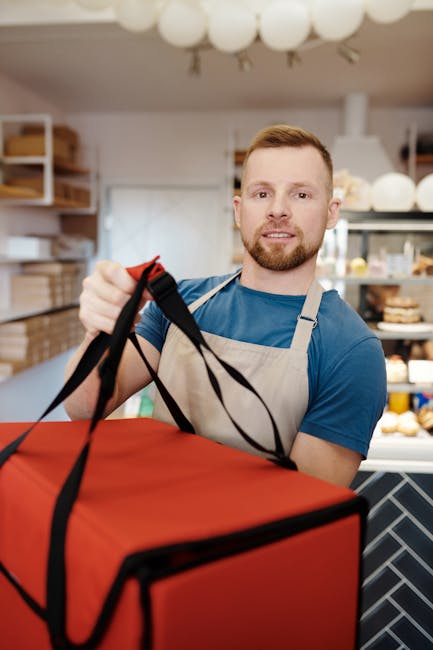 A young male florist with short, light brown hair and a beard, wearing a blue t-shirt and a beige apron, is smiling while handing over a large red floral delivery box with black straps. He is in a wel