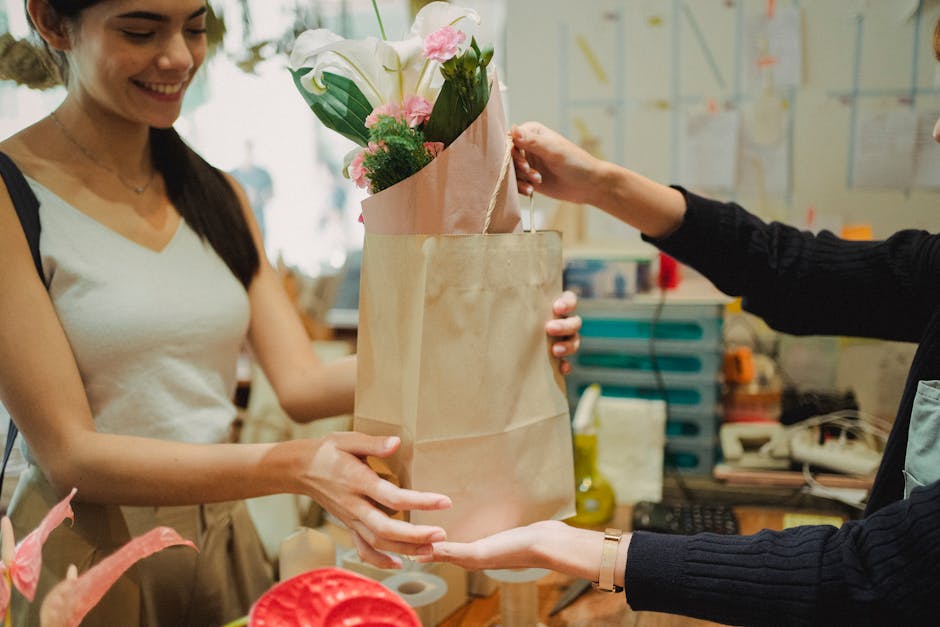 A woman with long dark hair smiling as she receives a bouquet of fresh flowers, which includes white lilies and pink blossoms, wrapped in pink tissue paper and paper wrapping, from another person at a