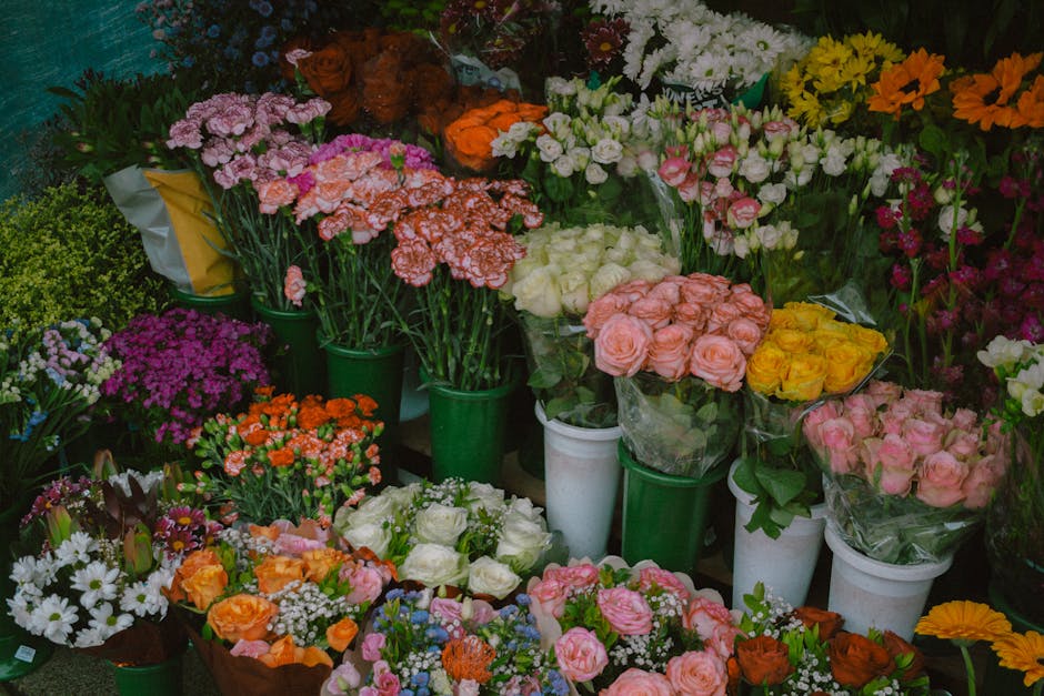 A vibrant display of various fresh flowers arranged in green and white buckets and wrapped bouquets, including pink roses, yellow roses, white roses, orange carnations, pink carnations, purple asters,