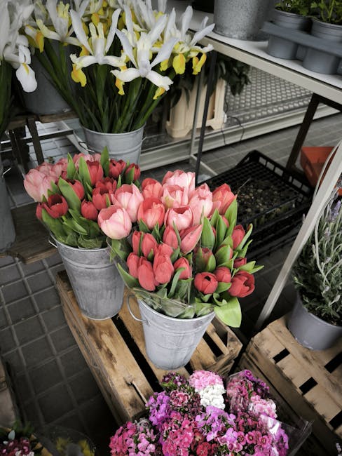 A floral display featuring two galvanized metal buckets of pink tulips with green stems and closed buds, arranged on a rustic wooden pallet. Behind them, a large bouquet of yellow and white irises wit