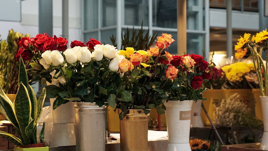 A variety of fresh flower bouquets displayed outdoors on a table, featuring red, white, yellow, and peach roses, arranged in metallic and ceramic vases. The roses have lush green leaves and their peta