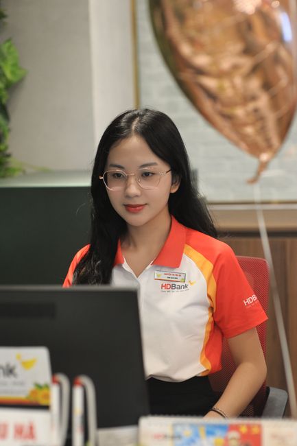 A young woman with long black hair and glasses is seated at a desk in an office environment, wearing a red, white, and yellow uniform with a name badge and the HDBank logo. She appears to be focused,