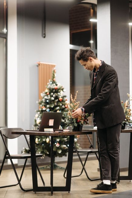 A young man with curly dark hair, dressed in a dark pinstripe suit and white shirt, arranging a floral gift on a table indoors. The bouquet features a mix of red, white, and green flowers, including r