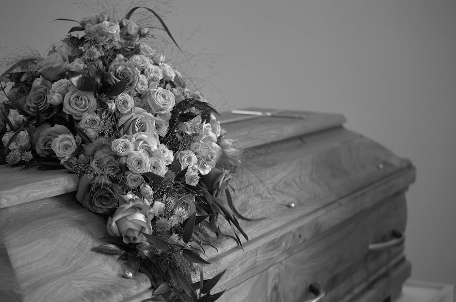 A large, meticulously arranged floral bouquet resting atop a polished wooden coffin. The bouquet features an assortment of fresh roses in various sizes, with some fully bloomed and others budding, pre