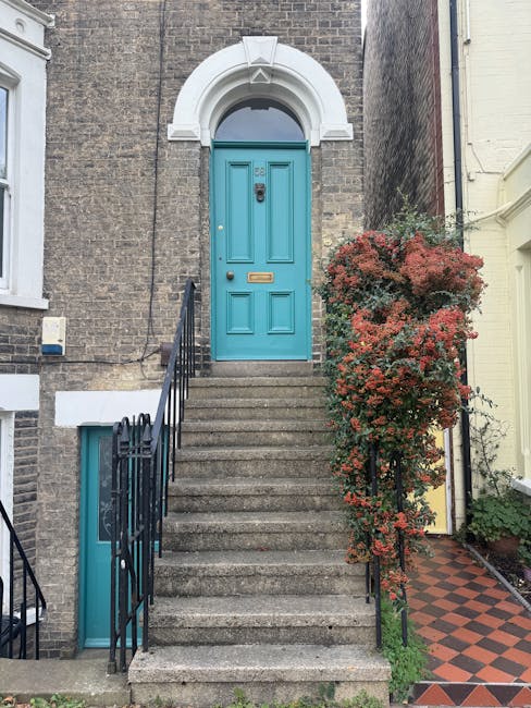 A vibrant turquoise front door set within a brick building, accessed by a wide concrete staircase with black metal railings on either side. To the right of the steps, a large flowering shrub with redd