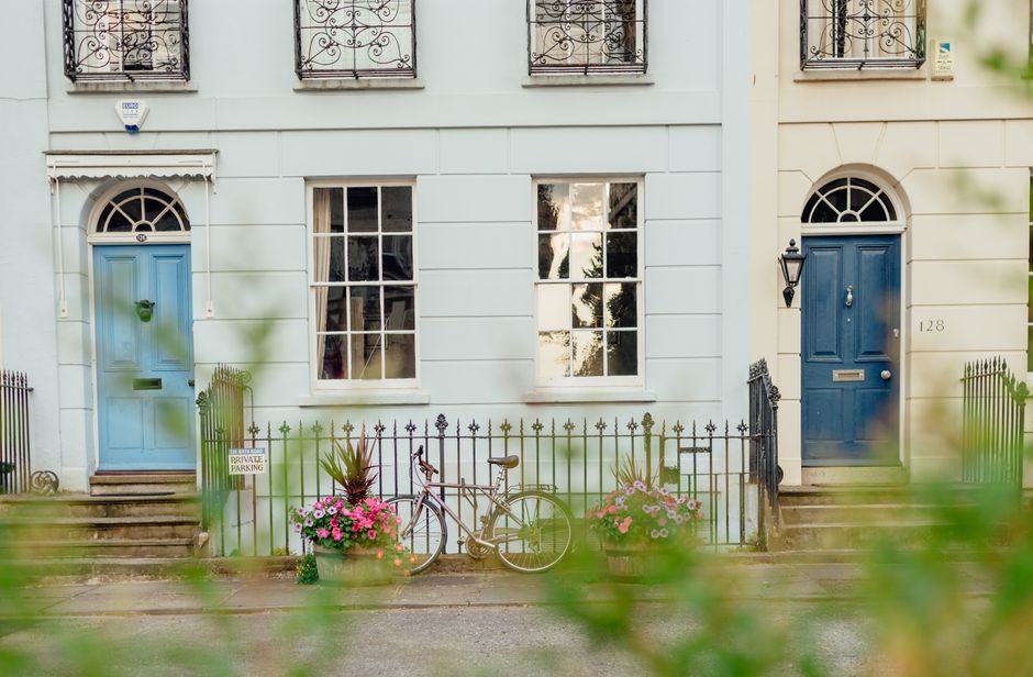A white facade of a residential building with two blue-painted front doors and large white-framed windows. In front of the building, a black wrought iron fence encloses small garden areas with two vib