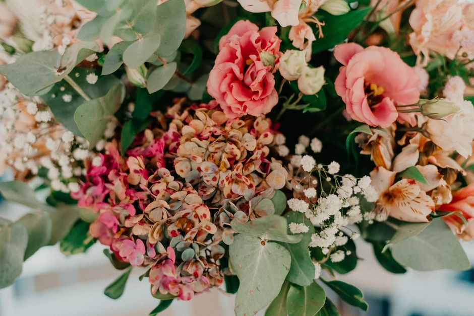 A close-up of a floral arrangement featuring a variety of fresh flowers and greenery. Prominent blooms include soft pink and peach-toned roses, clustered white baby's breath, and pink and brownish-gre