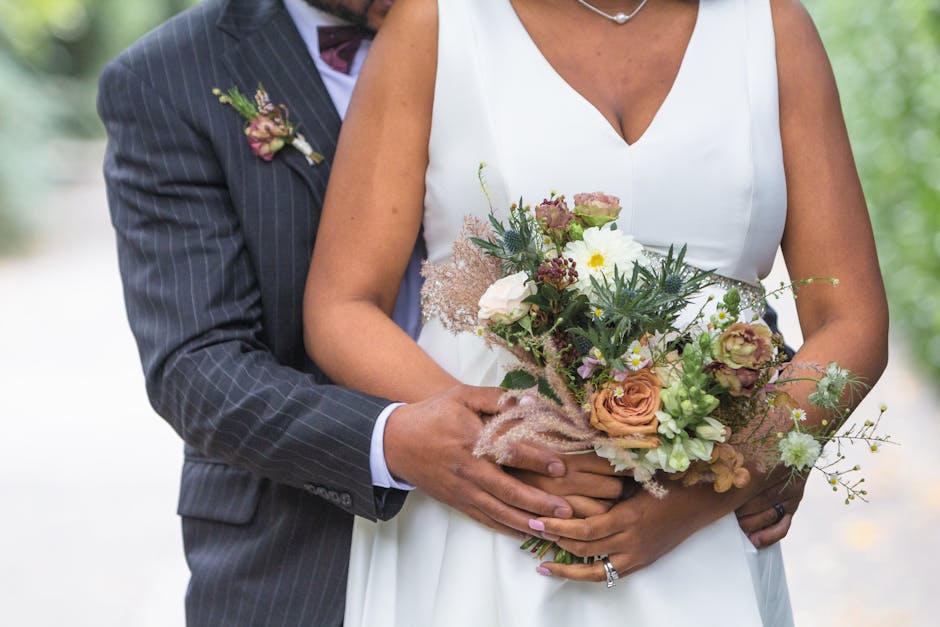 Close-up of a bride holding a floral bouquet featuring peach, white, and mauve roses, interspersed with green foliage and delicate filler flowers, wrapped in soft pink tissue paper. The bride's white 