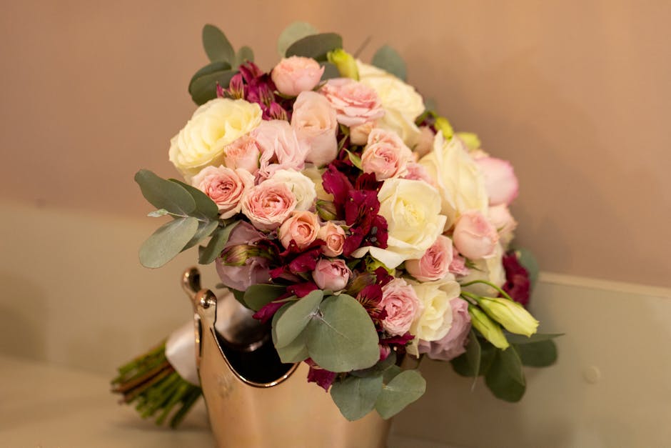 A close-up of a fresh floral bouquet featuring soft pink roses, creamy white lisianthus, and deep burgundy alstroemeria, accented with green eucalyptus leaves. The bouquet is arranged in a rounded, co