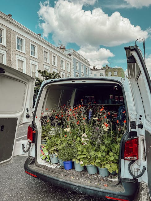 The open back of a white delivery van filled with potted flowering plants, including red poppies, white daisies, and green foliage. The plants are arranged in blue and gray pots, ready for floral arra