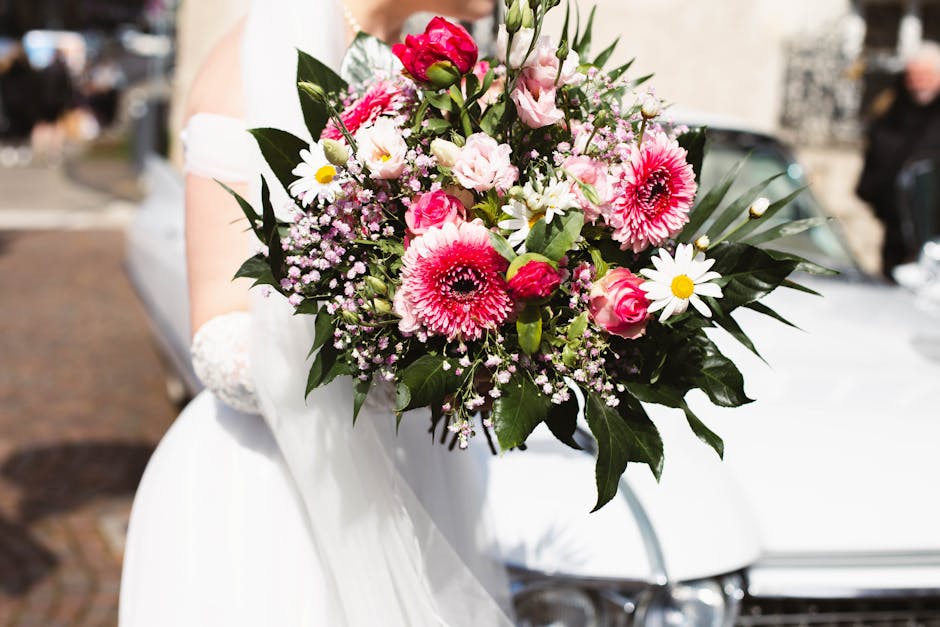 A person dressed in white holding a large, vibrant bouquet featuring pink gerbera daisies, pink and white roses, white daisies, and small pink filler flowers, all arranged with lush green foliage. The